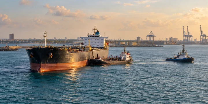 Tanker and two service craft off a low coast with container cranes in the distance