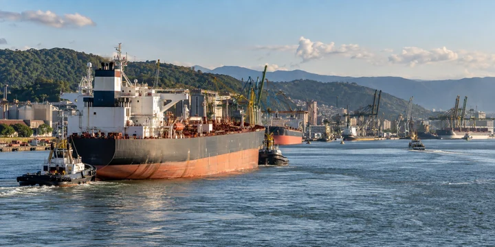 Tanker guided by small craft in a harbor below wooded hills and cranes